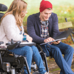 A man in a red hat looking at a laptop with a woman in a wheelchair.