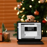 A laptop featuring an LMS-specific How to Fix video sits on a desk in front of a holiday tree, with wrapped presents on one side and a mug on the other. A teddy bear hangs off the laptop.