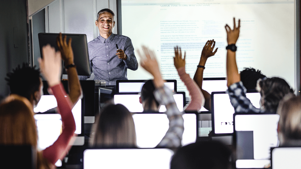 An instructor stands in front of a classroom full of students raising their hands.